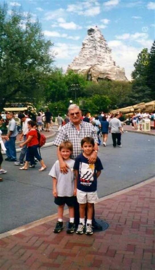 Two children pose with an adult at a busy amusement park on a sunny day.