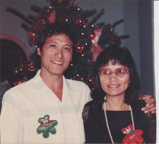 Couple poses happily in front of a Christmas tree adorned with lights and decorations.