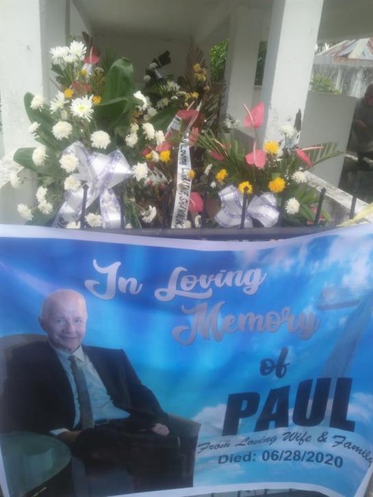 Flowers and a banner pay tribute to Paul in a peaceful garden, honoring his memory with love.