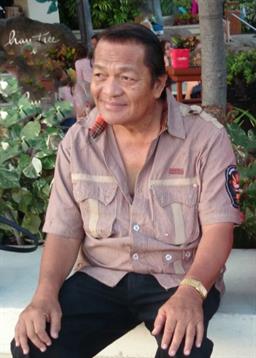 A relaxed senior man smiles while seated outside surrounded by lush plants in afternoon light.