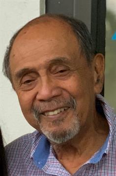 Elderly man smiles warmly while seated outdoors, surrounded by green plants and flowers.