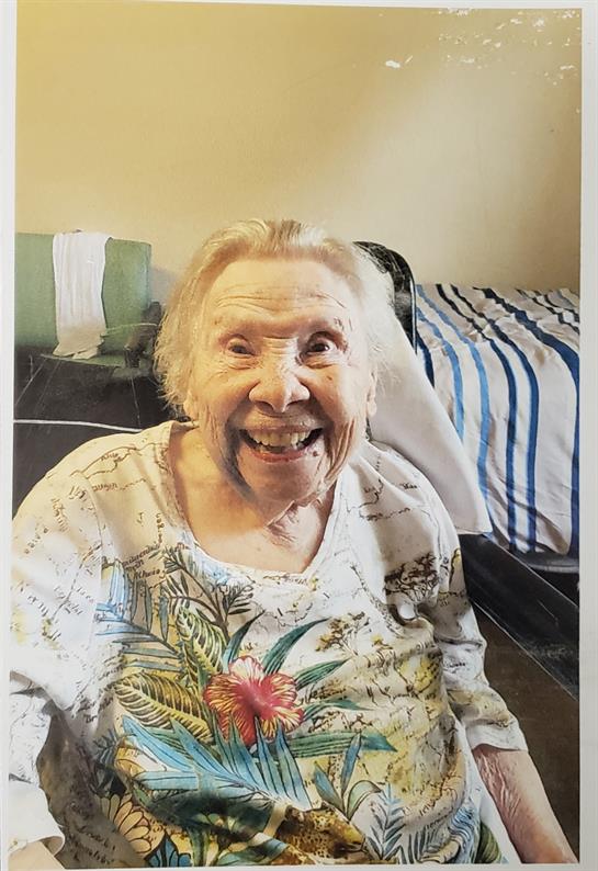 An elderly woman sits in a chair, grinning widely in a bright, inviting room with simple decor.
