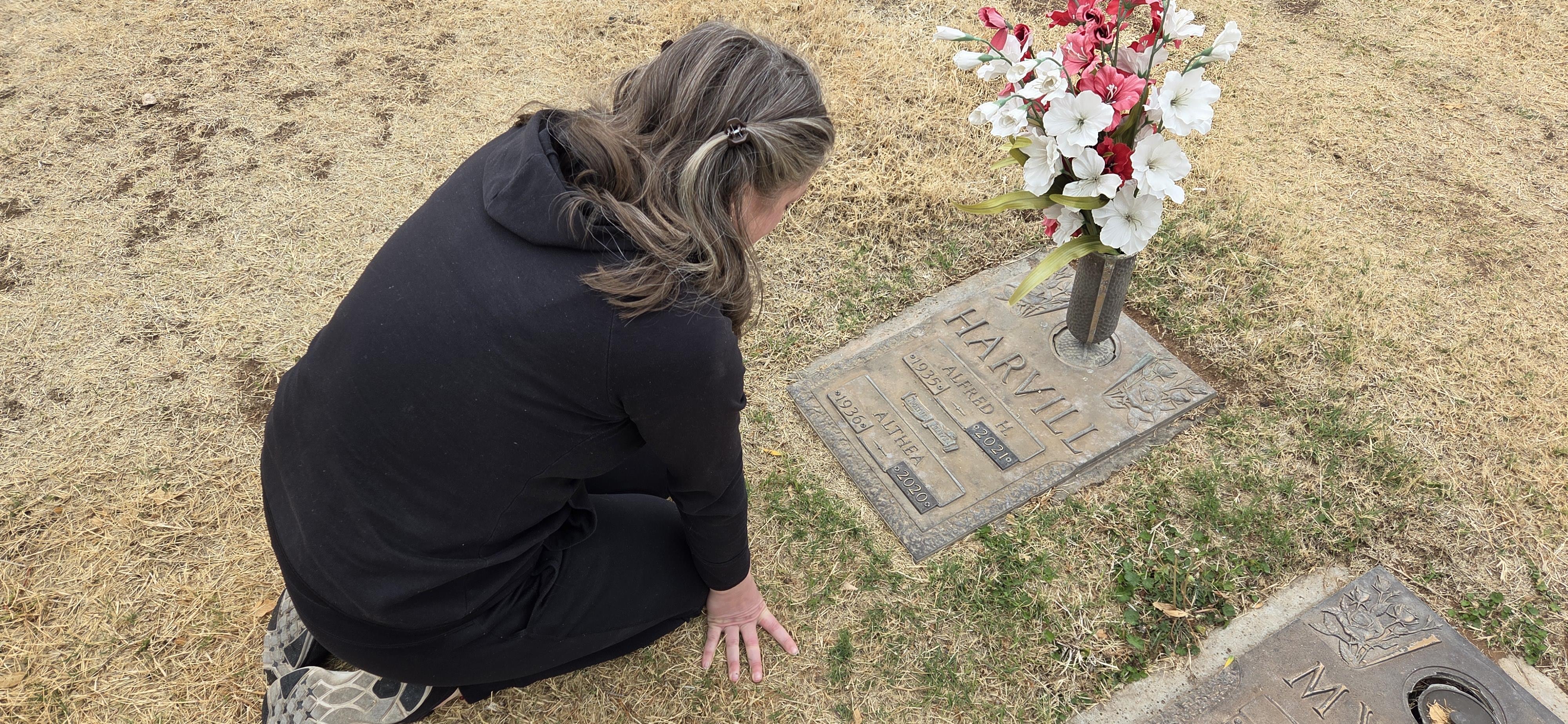 A woman kneels in a cemetery, reflecting by a grave adorned with flowers in a moment of remembrance.