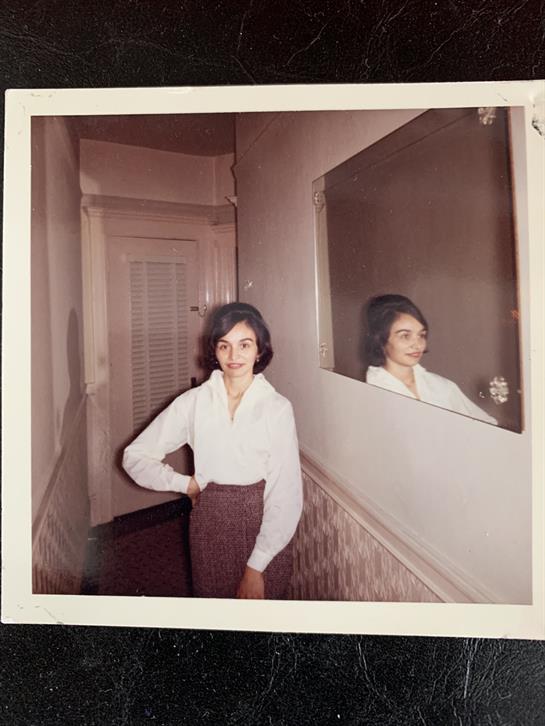 A woman stands in a hallway, smiling and posing beside a mirrored wall.