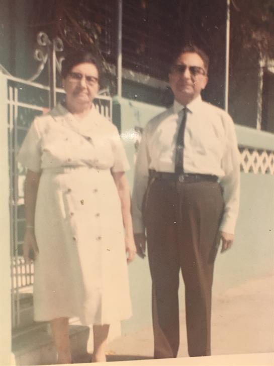 A couple poses for a portrait outside a building, wearing vintage attire on a warm day.