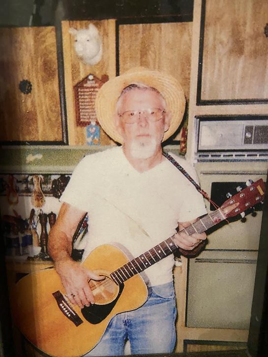 Elderly musician wearing straw hat enjoys playing guitar in a cozy kitchen environment.