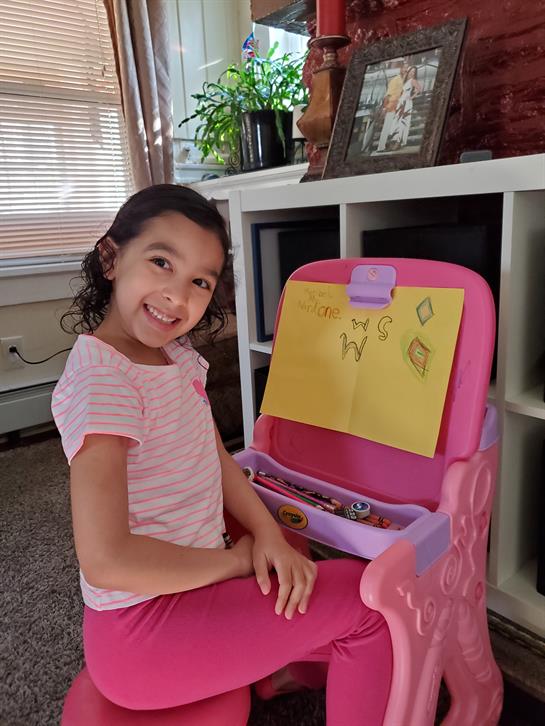 Young girl smiles while crafting at a colorful desk filled with art materials in her home.