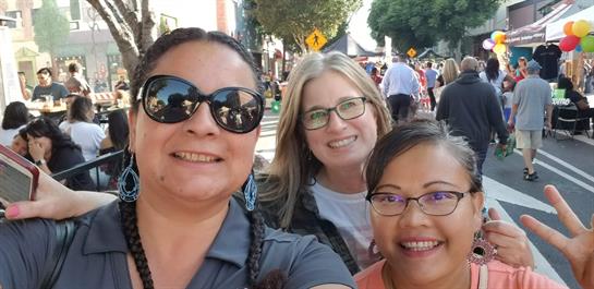 Three women take a cheerful selfie during a vibrant street festival filled with activity.