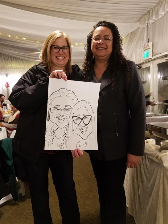 Two women smile as they hold up a caricature artwork of themselves during a party.