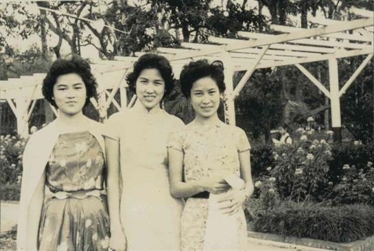 Three women stand side by side in a lush garden, dressed elegantly in traditional attire, smiling.