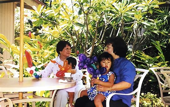 Two adults enjoy conversation while a child holds colorful balloon creations amid greenery.