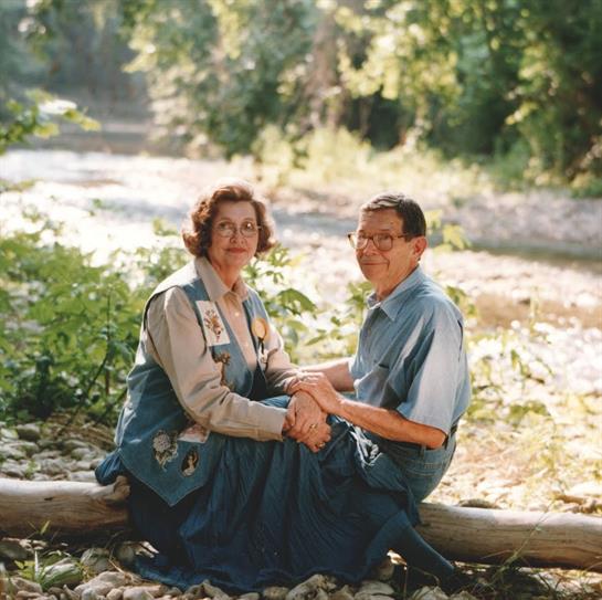 Two people sit together on a log by a river, surrounded by lush greenery and tranquility.