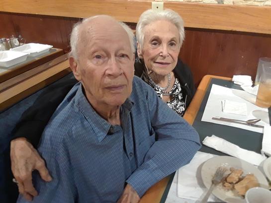 Two elderly individuals sit closely together at a table, enjoying a meal in a relaxed atmosphere.