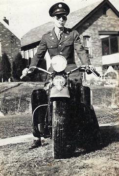 A motorcyclist in uniform proudly sits on a vintage bike near stone buildings, showcasing history.