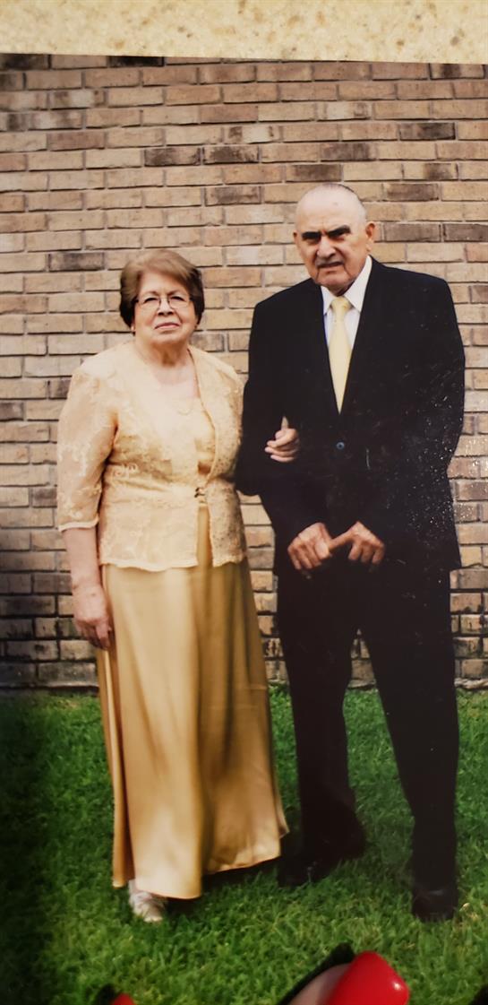 An elderly couple stands hand in hand, dressed formally, smiling in front of a brick wall.