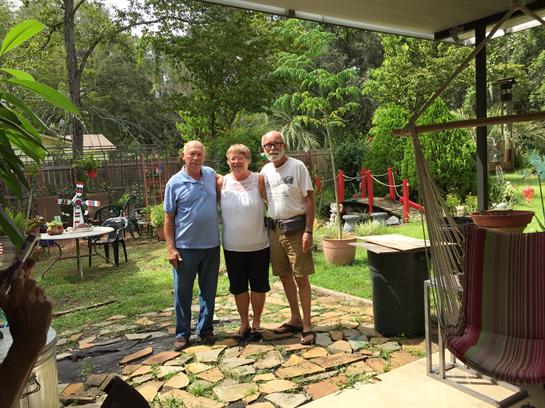 Three friends stand smiling in a vibrant garden surrounded by greenery and outdoor furniture.