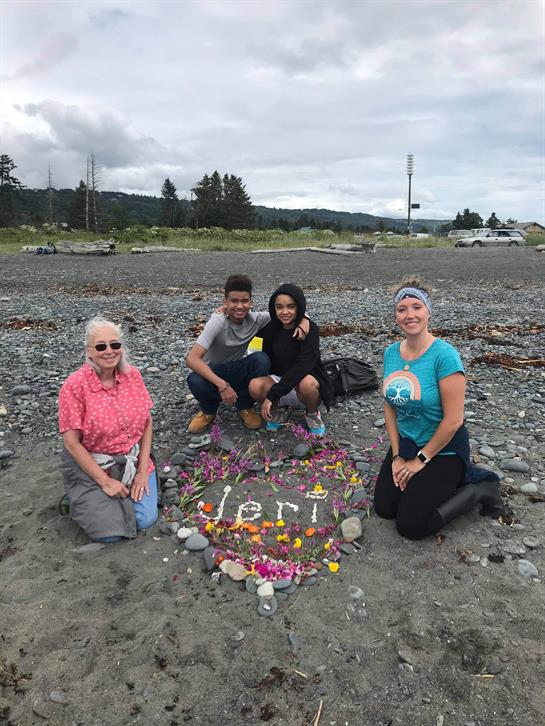 Four individuals on a beach arrange colorful stones to form a heartfelt design for someone special.