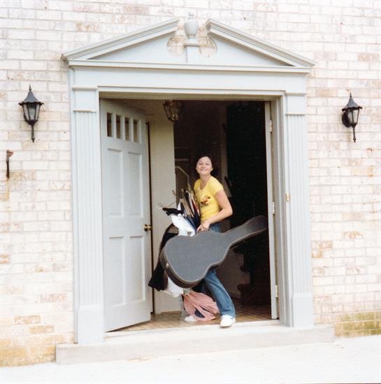 A man with a guitar and backpack steps out of a house dressed casually and smiling warmly.