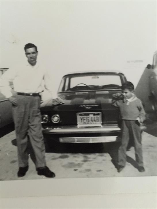A man and a young boy stand next to a classic car, enjoying a sunny day in a casual outdoor setting.