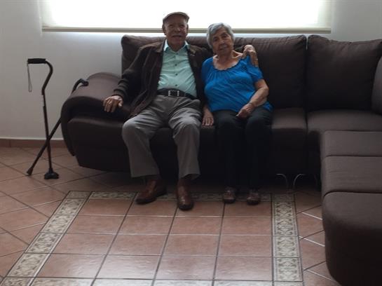 An elderly couple sits closely together on a dark sofa in a bright, welcoming living room.