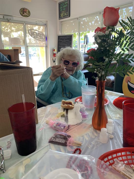 An elderly woman in sunglasses savors her dessert while seated at a cafe table adorned with flowers.