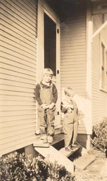 Two children dressed in vintage clothing play outside a house, enjoying a sunny day together.