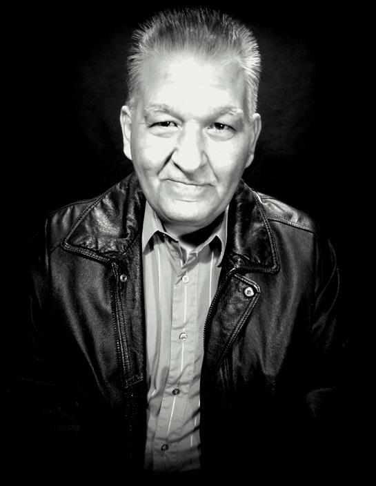 Man with gray hair smiles while sitting in a studio, wearing a leather jacket and shirt.