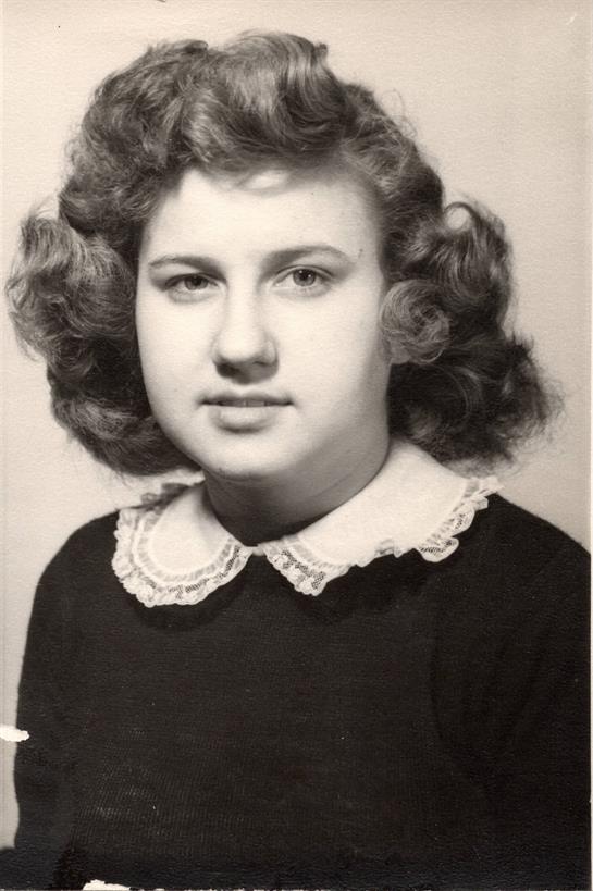 A young woman with curly hair wears a dark dress with a white collar, posing indoors.