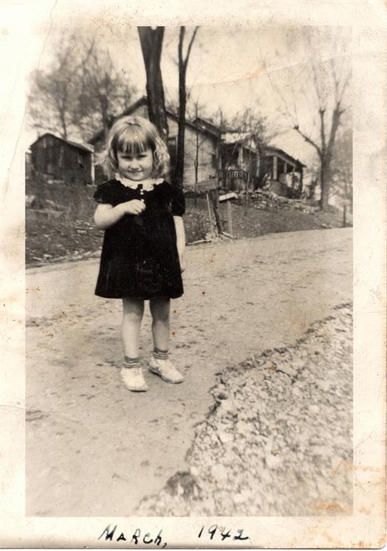 A smiling blonde girl stands on a dirt road with rustic houses behind her.