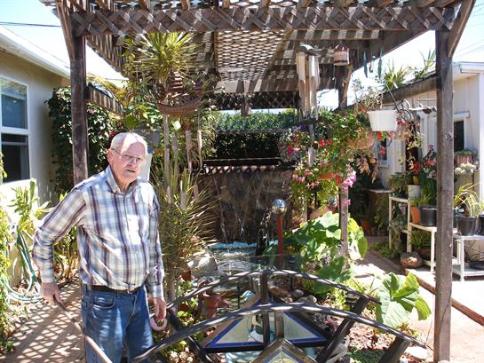 An elderly man enjoys a sunny afternoon in a lush garden filled with plants and flowers.