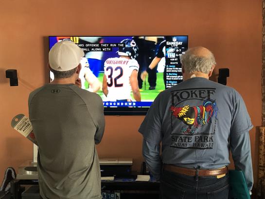 Two men stand together focusing on a football game displayed on the television screen.