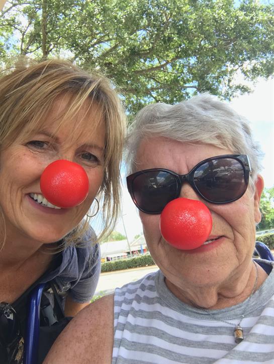 Two women share smiles while wearing bright red clown noses on a sunny day outdoors.