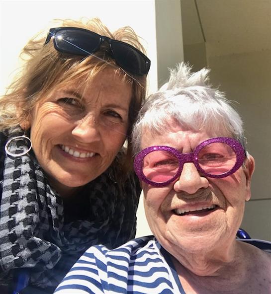 Two women are joyfully smiling together outside, wearing sunglasses, and sharing a special moment.