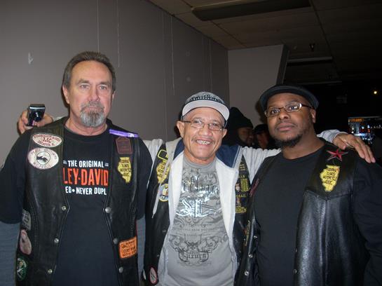 Three men wearing leather motorcycle club jackets are posing together in a lively venue.