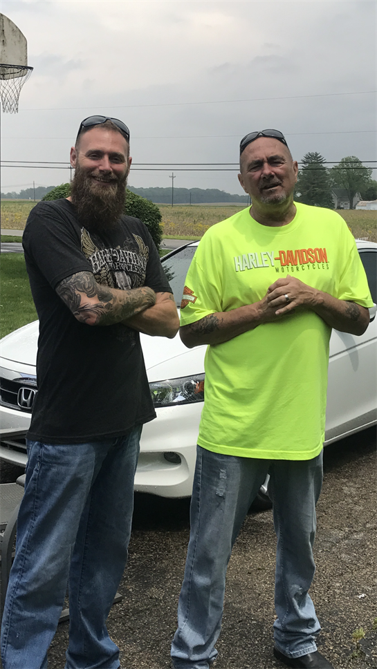 Two men pose confidently beside a white vehicle in a rural setting, enjoying a sunny day.