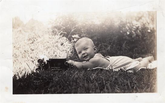 A baby is lying on the grass, smiling at a toy train while enjoying a sunny day outdoors.