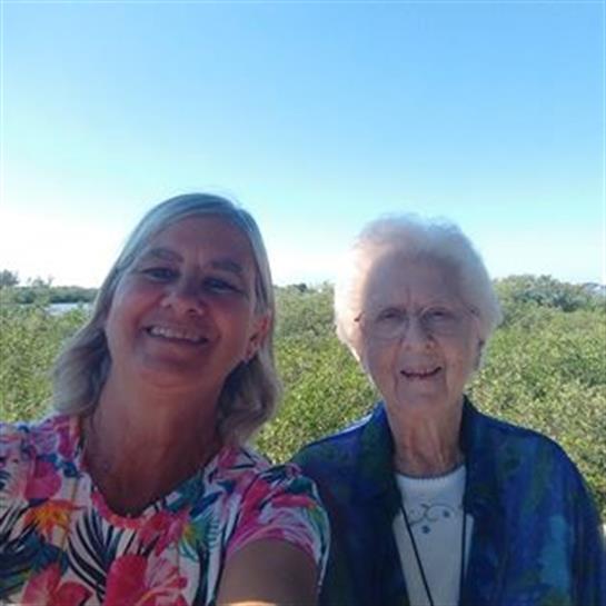 Two women are taking a cheerful selfie outdoors, surrounded by lush greenery and a bright blue sky.