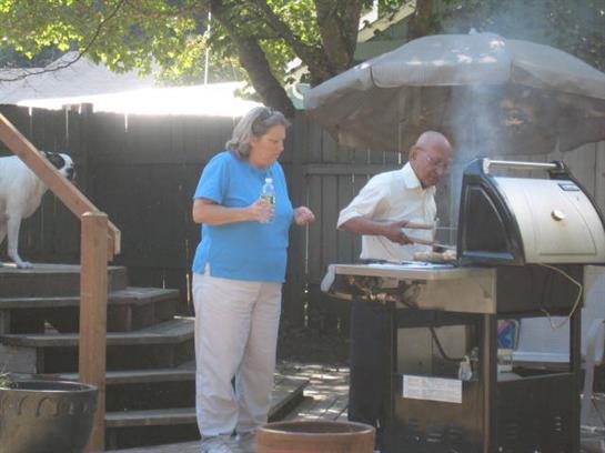 Two people grill food together in a backyard while enjoying a sunny outdoor day.