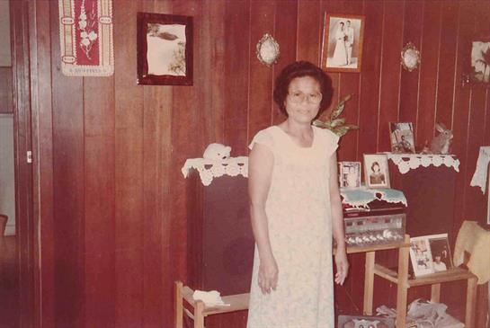 An elderly woman poses in a cozy wooden room filled with family pictures and traditional decor.
