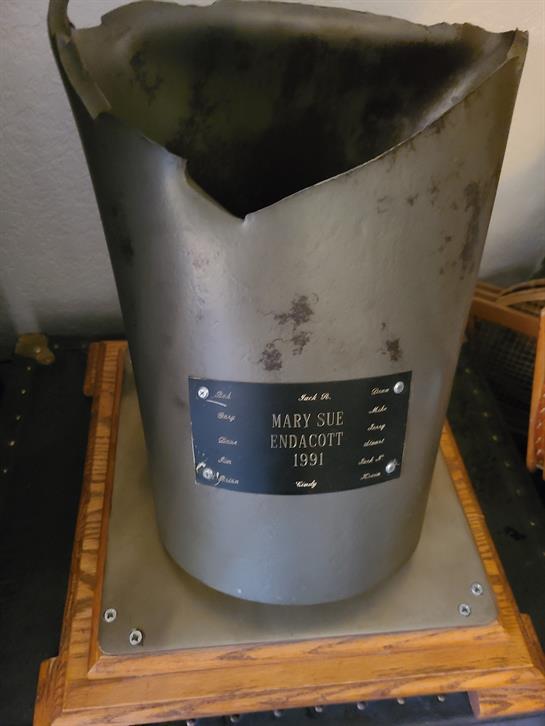 A damaged metal artifact sits on a wooden base, showcasing its history through a plaque nearby.