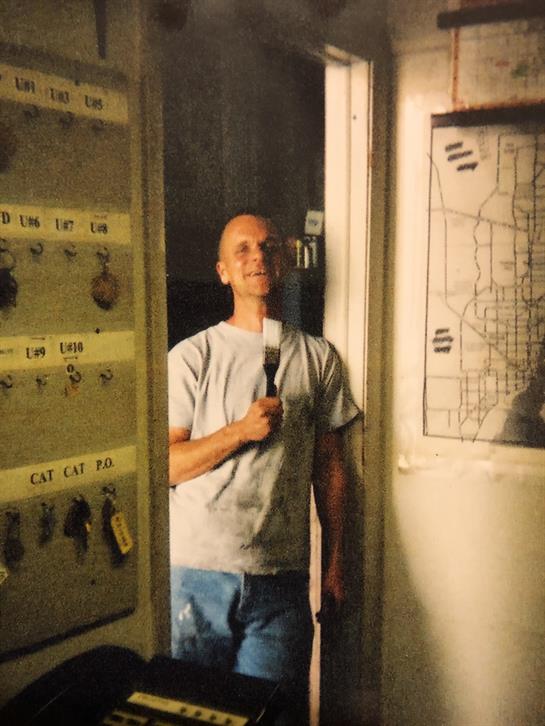A skilled worker smiles while holding a paintbrush in a workshop filled with tools and plans.