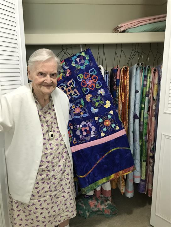 A smiling elderly woman stands by a closet showcasing beautiful quilts and vibrant fabrics.