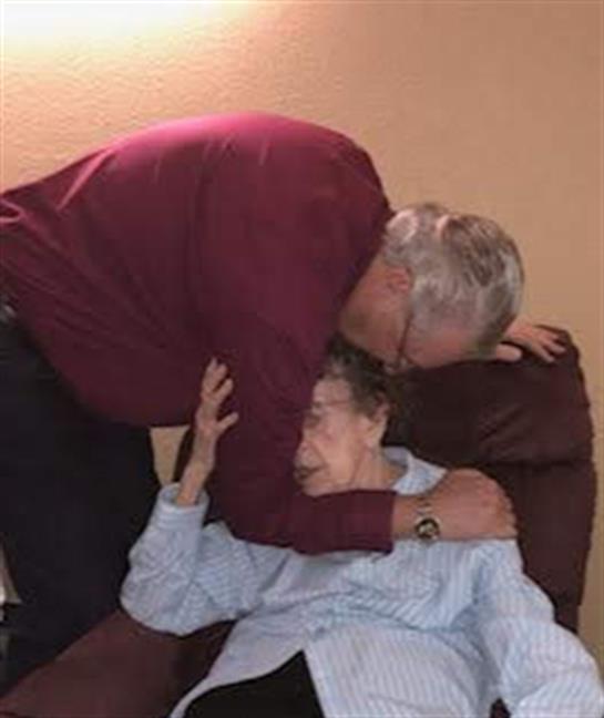 A heartfelt moment between a man and an elderly woman in a comfortable chair shows deep affection.