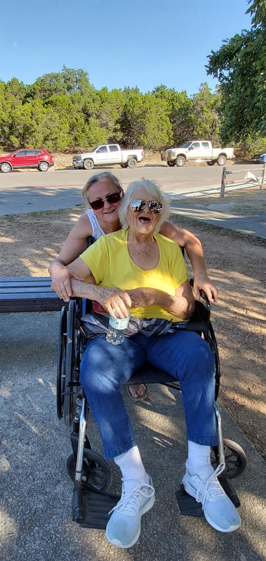 Two women share a joyful moment in a park, with one woman in a wheelchair smiling in the sun.