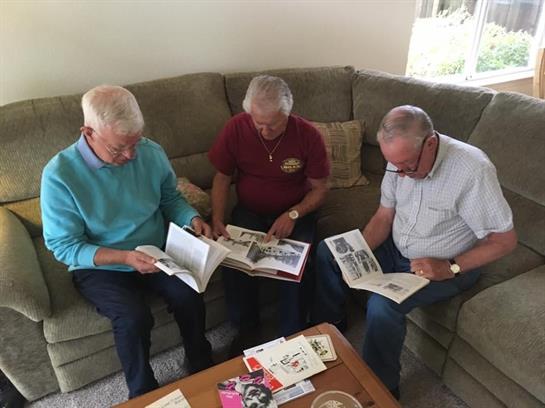 Three older gentlemen share stories while looking through magazines on a couch.