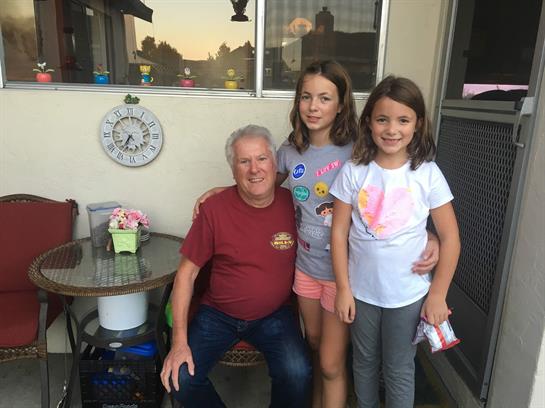 Two young girls stand beside an older man, all smiling happily in a cozy outdoor area during sunset.