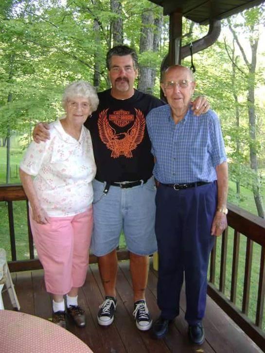 Two elderly individuals and a man smile for a group picture on a porch in the warm sunlight.