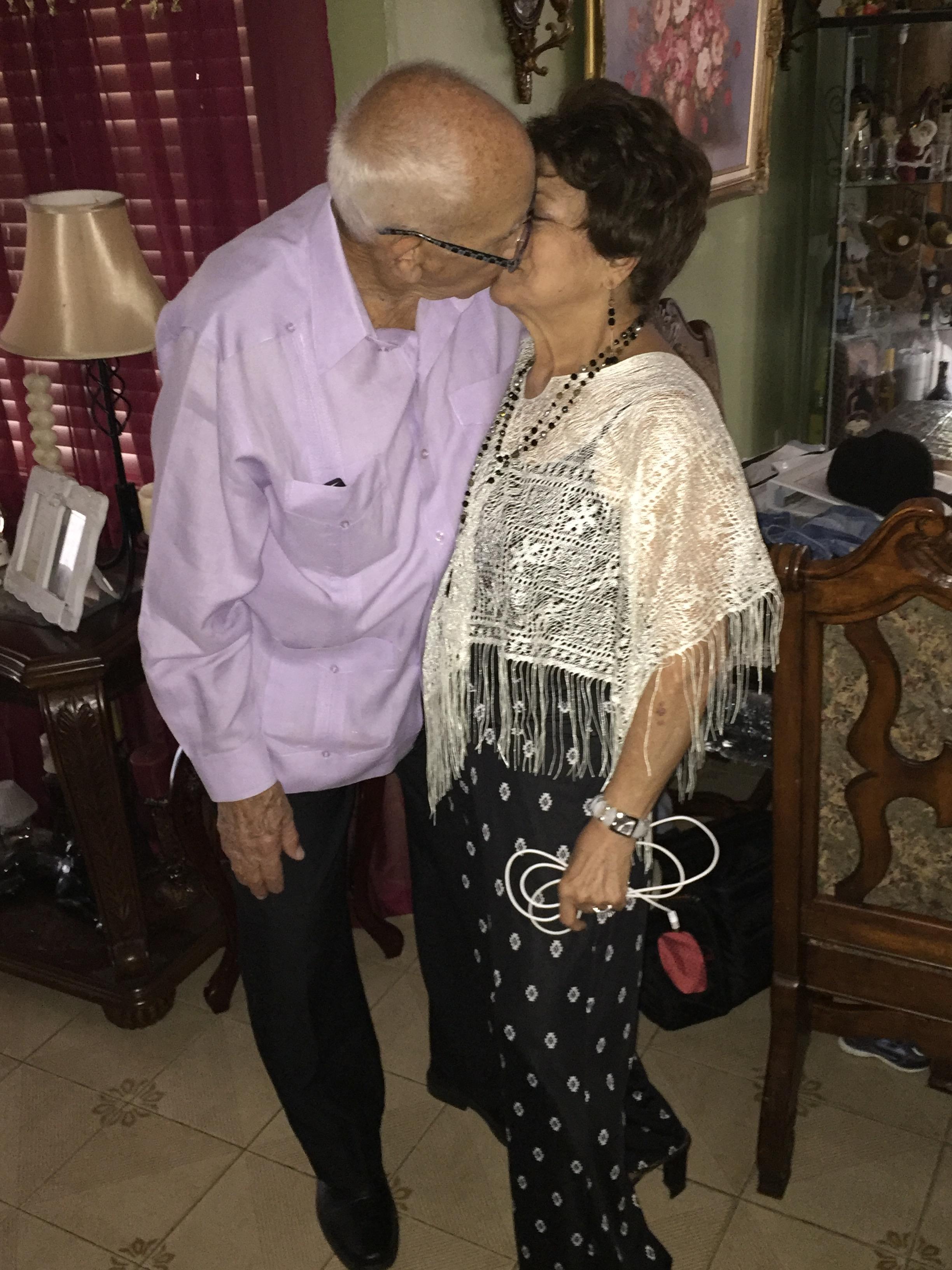 An elderly couple is smiling and leaning toward each other in a living room.