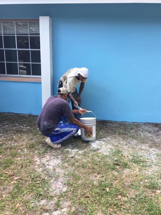Workers carefully paint a blue wall, focusing on details while using a bucket of paint outdoors.