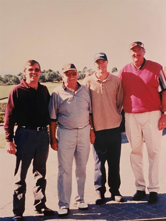 Four friends pose together on a golf course during a clear afternoon, smiling and relaxed.